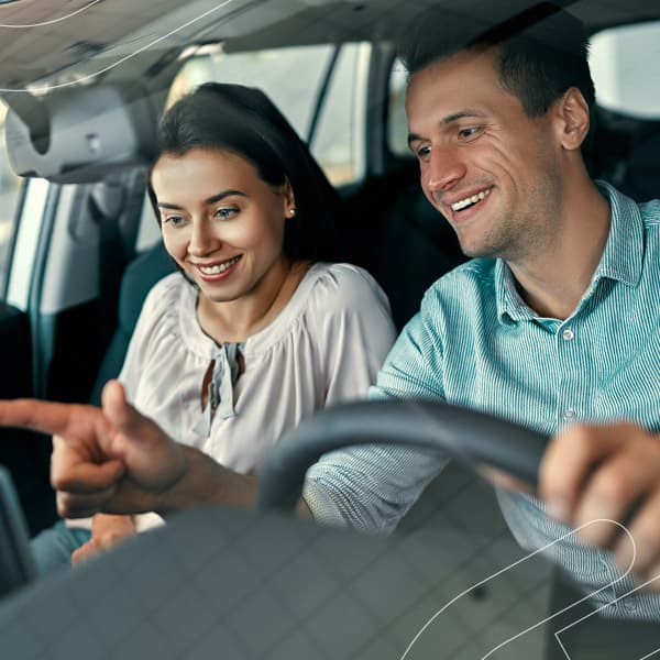 Casal sorrindo dentro de um carro enquanto o homem aponta para o painel do carro